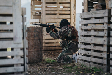 Fototapeta premium A camouflaged player crouches behind pallets, aiming a paintball marker in a staged outdoor course.