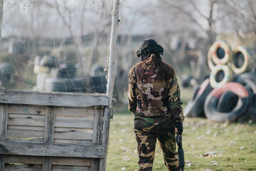 Fototapeta premium A camouflaged player stands with a marker, facing a paintball field with tires and netting.