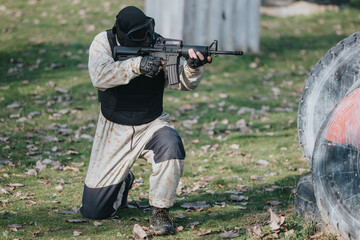 Fototapeta premium A person wearing a helmet, vest and camo kneels and aims a rifle on a paintball field. The scene shows a focused tactical player in protective gear amid an outdoor park setting.