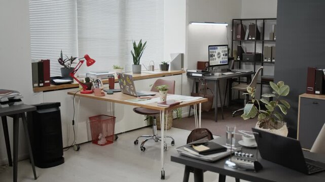 Interior of modern office featuring wooden desks with laptops, documents and notebooks, also grey cabinet, file folders and potted plants on windowsill, trash bins