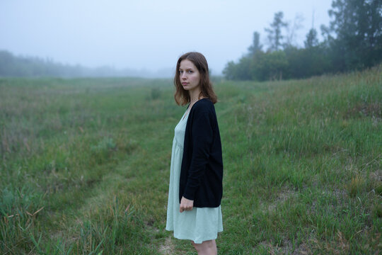 Woman in dress standing in foggy summer meadow with forest in morning
