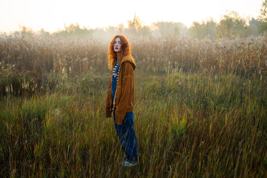 Red haired woman in orange hoodie standing in foggy sunlit field