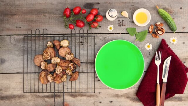 A grill with baked potatoes and zucchini was placed on an old wooden table. Cucumber, tomatoes, spinach, walnut, garlic clove, pepper, oil, and salt were on the table.