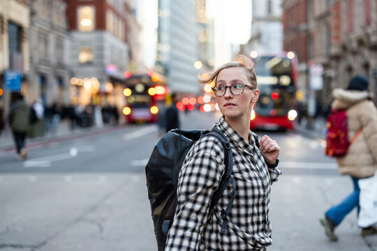 Hipster commuter with backpack crossing busy city street at dusk