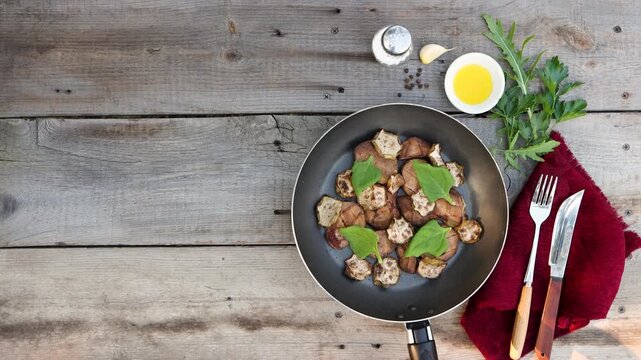 A frying pan with fried potatoes and zucchini with spinach was placed on an old wooden table. Parsley, arugula, a clove of garlic, peppercorns, and vegetable oil were on the table.
