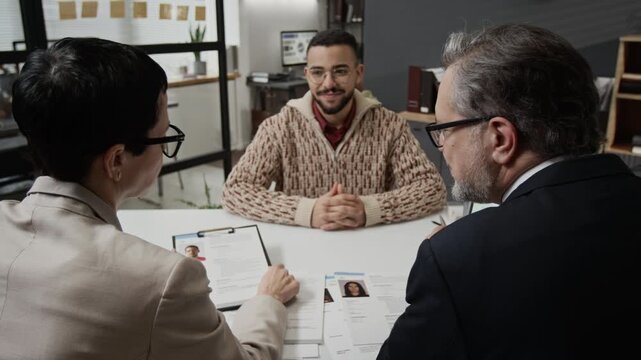 Zoom in over the shoulder view of two middle-aged recruiters in office wear offering job to gen Z applicant and giving him form for signing after conducting interview at white table
