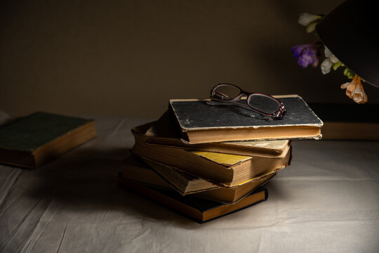 Stack of old books with reading glasses illuminated by desk lamp, cozy low light study still life.