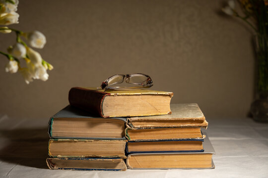 Stack of old books with reading glasses illuminated by desk lamp, cozy low light study still life.