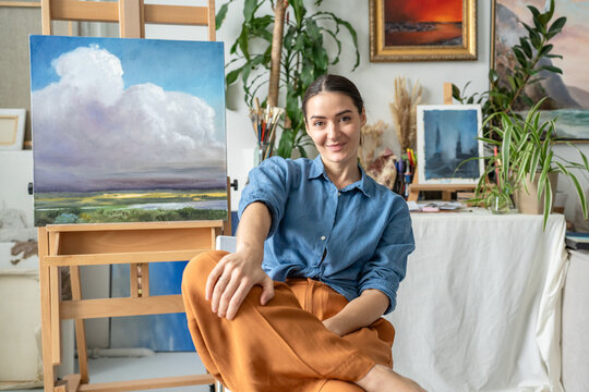 Cheerful positive woman artist posing in studio workshop surrounded by paintings and artworks. Smiling confident female painter sits beside painting display on easel, creative art hobby and therapy.