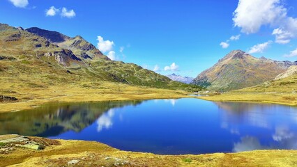 Fototapeta premium A tranquil lake mirrors rugged hills and towering mountains beneath bright skies. Green and brown terrain blends with clouds, creating a peaceful scene marked by vivid natural colors. 