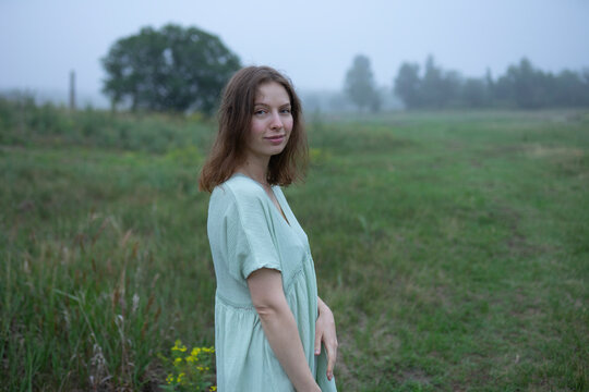 Woman in green dress standing in foggy summer field looking at camera