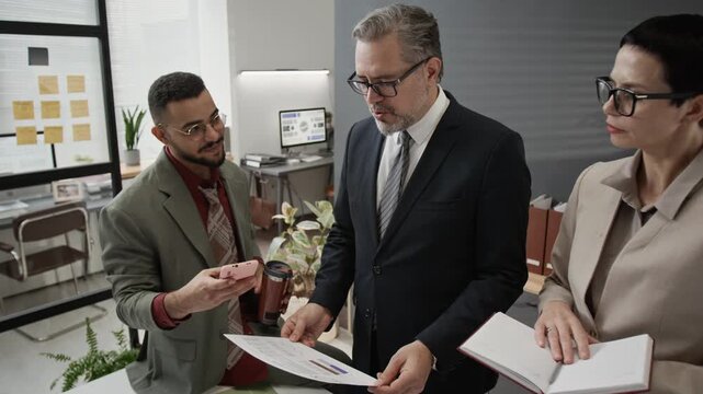 Positive zoomer taking selfies with two middle-aged colleagues during group discussion in office, they smiling but getting back to business