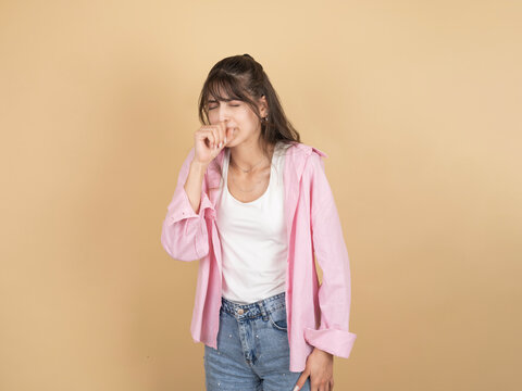 A young woman in a pink shirt coughs into her fist with her eyes closed, appearing unwell. She stands against a neutral beige background, representing symptoms of illness like a cold or flu.