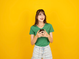 A smiling young woman in a green t-shirt looks sideways while holding a smartphone. She stands against a solid yellow background, portraying a happy reaction to a message or notification. © Designerant