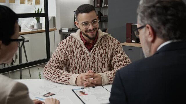 Over the shoulder zoom out shot of two mature colleagues interviewing positive young man applying for job in modern office