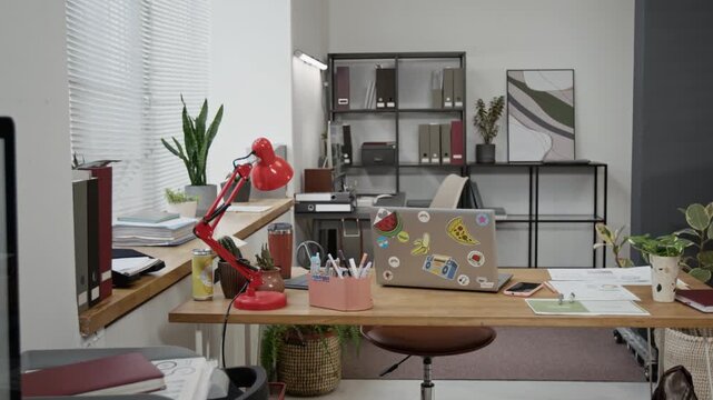 No people view of work table with laptop, desk lamp, thermos, documents, small plants, yellow can, notebook and smartphone, also black rack with file folders in background