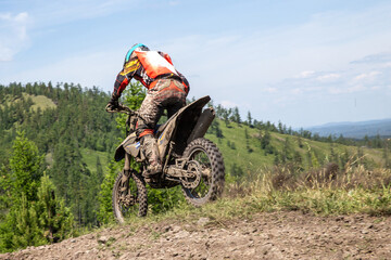 Rear view enduro rider on heavily mud-covered dirt bike performing wheelie on rocky hilltop trail © Real Sports Photos