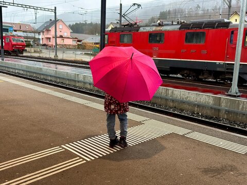 Switzerland &ndash; February 9, 2026: Child standing with umbrella at train station platform near red SBB locomotive during rainy weather.
