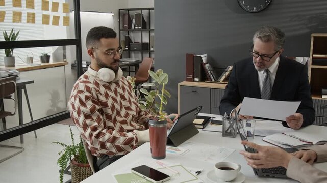 Two mature business people in formalwear working at office desk, while zoomer having drink, listening to music with headphones, squeezing hand expander and studying data on digital tablet