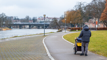 A man walks along a riverside path with a bike trailer stroller in Germany. The scene is set in a park with trees and a bridge in the background.