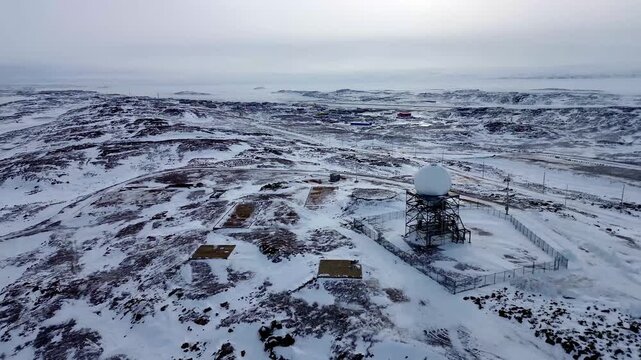 Aerial drone push-in toward a remote North Warning System radar dome. High-tech surveillance infrastructure isolated in the vast, snow-covered Arctic tundra near Iqaluit.