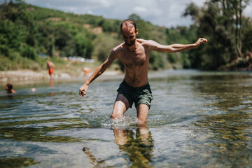Fototapeta premium Man wades through shallow river water, splashing with outstretched arms. The sunny outdoor scene captures a casual river moment by nature.
