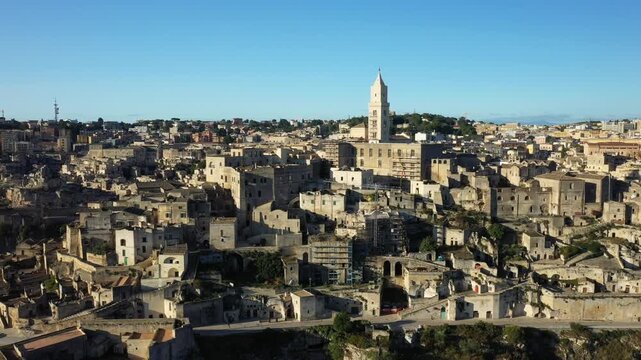 Aerial view of the historic stone buildings and iconic cathedral of Matera, Italy, under a clear blue sky. The unique architecture and dramatic landscape highlight the charm of this ancient city.