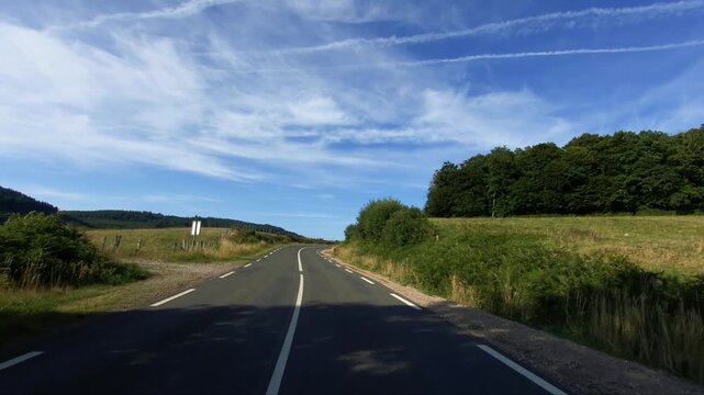 Curving rural road in the Morvan region, bordered by green vegetation and fields under a bright blue sky with wispy clouds.