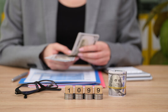 A business professional sorts through cash while focusing on 1099 tax paperwork at a neat desk. Nearby, glasses rest on stacks of coins, highlighting a busy financial day.
