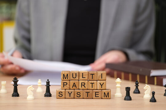 On a warm wooden desk, chess pieces surround blocks spelling MULTI PARTY SYSTEM as a person engages in a thoughtful political discussion, papers in hand, lost in strategy.
