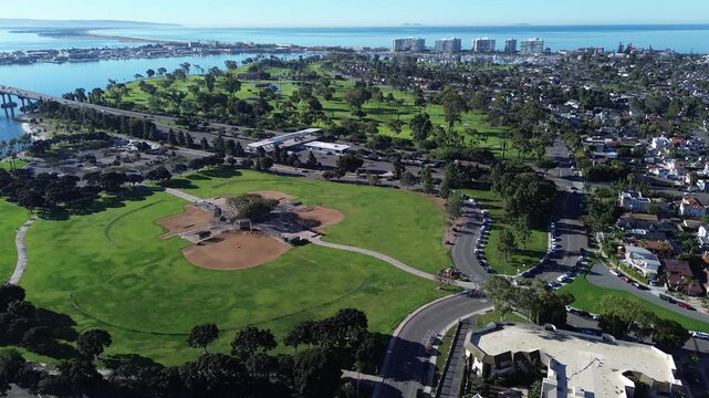 Waterfront recreation zone spans Tidelands Park, Glorietta Blvd, Coronado Bridge corridor. Bright turf, ballfields sport complex, patterned tree canopies, reflective bay tones, Coronado, CA