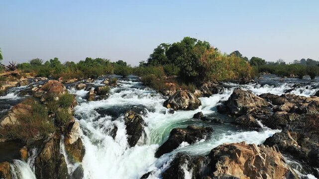 Scenic view of Tat Somphamit (Li Phi) Waterfalls cascading over rocks surrounded by lush greenery at sunrise in Laos. Powerful river rapids and natural beauty in a tropical landscape.