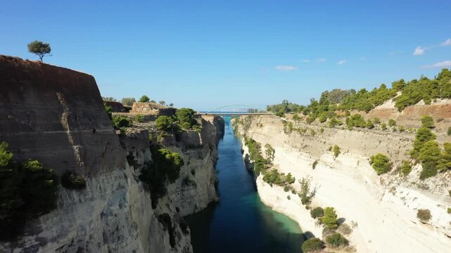 Aerial view of the Corinth Canal in Greece, featuring steep limestone cliffs, clear blue water, and a bridge spanning the canal under a clear sky.