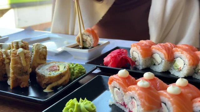 Beautiful attractive pretty girl in a good mood eating sushi in a cafe. Dip sushi in soy sauce, a serving of various fried hot sushi, a Philadelphia girl eating sushi with chopsticks