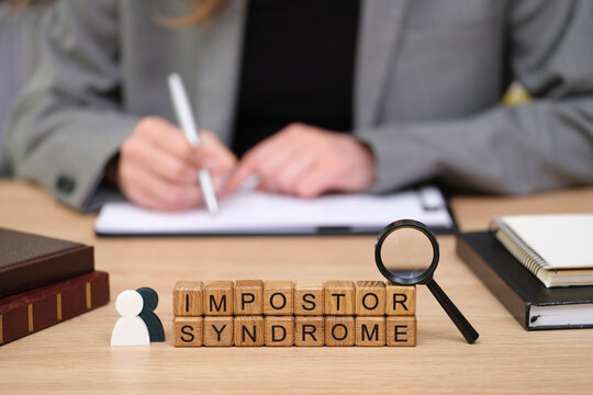 In a contemporary office, a person is focused on writing notes while blocks spelling out impostor syndrome sit nearby, illustrating the struggle with self-doubt and confidence.