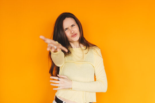 Skeptical young woman pointing at camera with disbelief on orange background, annoyed girl looking with contempt, judging and negative emotions concept, studio portrait