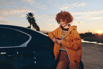 A smiling woman in a rainbow sweater and orange jacket stands next to a black car outdoors at sunset while using a smartphone. The woman enjoys a casual lifestyle moment. © SHOTPRIME STUDIO