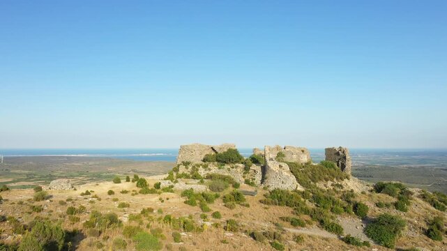 Ancient stone ruins of Chateau de Opoul Perillos overlooking a scenic rural landscape under a clear blue sky. Wide view of the historic fortress surrounded by dry grass and distant coastline.