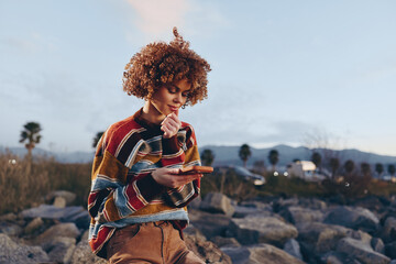 Young woman with curly hair wearing rainbow sweater smiles while using smartphone outdoors near...
