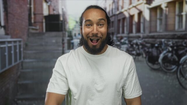 Man in white t shirt points finger at camera on narrow city street with rows of bicycles and brick building; surprise engagement invitation.