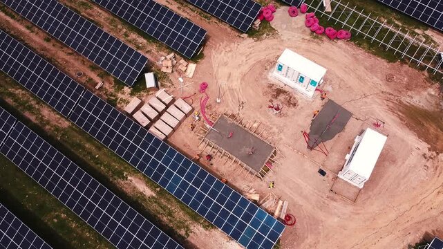 Cinematic drone roll shot above a solar farm construction site, revealing transport and staging of solar panels and mounting frames beside a dirt road and machinery.
