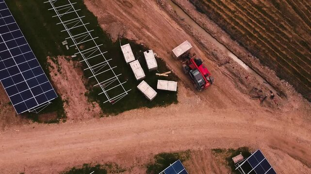 Cinematic drone roll shot above a solar farm construction site, revealing transport and staging of solar panels and mounting frames beside a dirt road and machinery.