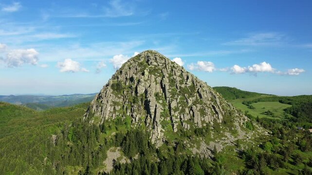 Aerial view of the iconic Mont de Gerbier de Jonc, a rocky volcanic mountain surrounded by lush green forest in Ardeche, France under a clear blue sky.
