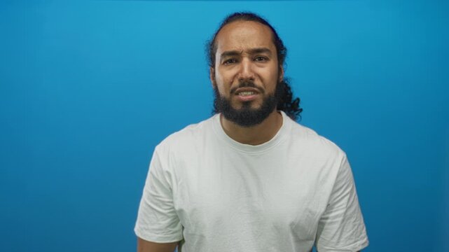 Man with beard squinting and mouth open wearing white t shirt in studio with blue backdrop; confusion.
