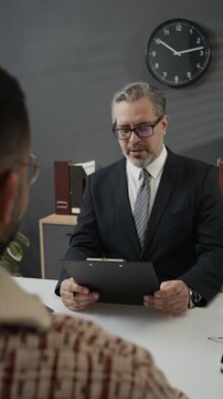 Vertical over the shoulder view of confident grey-haired HR specialist in formalwear interviewing young man and taking notes holding clipboard in office