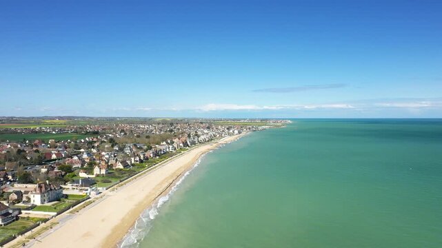 Aerial drone shot of Sword Beach and the coastal town of Hermanville sur Mer in Normandy, France, with clear blue sky and turquoise water. Picturesque seaside landscape with residential houses lining