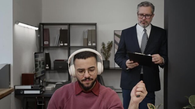 Gen Z guy with headphones drinking from insulated tumbler and squeezing hand expander while working on project in front of laptop, mature man holding business papers looking at him with confusion