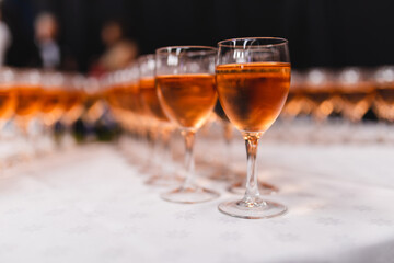 Fototapeta premium Rows of elegant wine glasses filled with chilled rose wine or amber cider on a white tablecloth. Shallow depth of field with a blurred background of a festive event, celebration, or gala dinner.