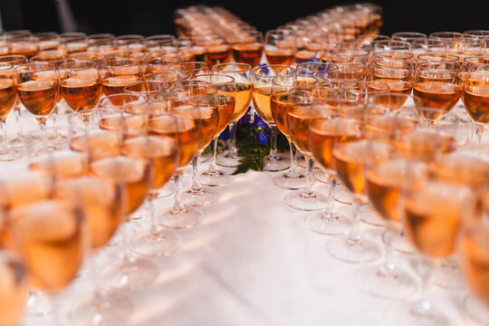 Rows of elegant wine glasses filled with rose wine or champagne on a white tablecloth. Symmetrical catering arrangement for a wedding, luxury gala, or festive celebration with warm lighting.