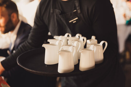 Professional waiter in a black apron carrying a round tray filled with several white ceramic milk pitchers or creamers, serving guests at a catered event, banquet, wedding, or elegant restaurant.